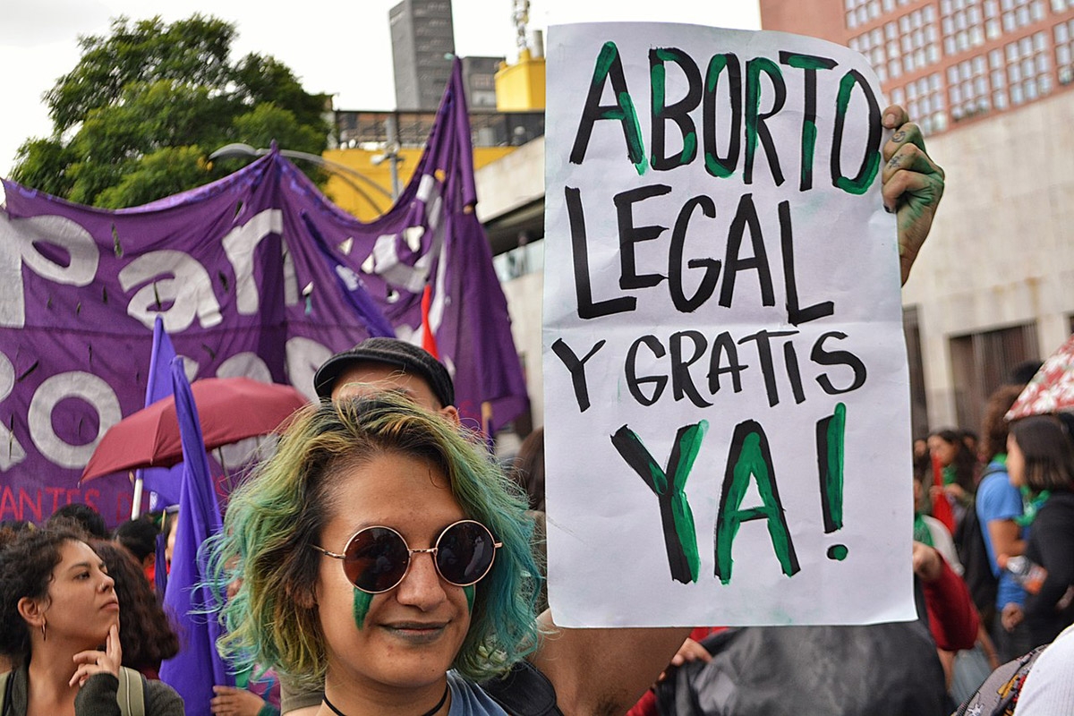 Bild einer Demonstration ein Schild im Vordergrund trägt den Text "Aborto legal y gratis ya!"
