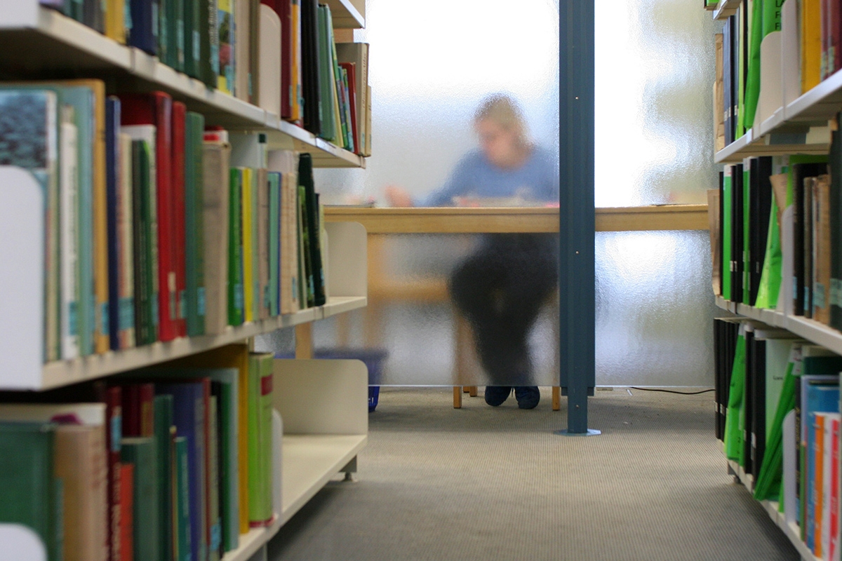 Person sitzt am Ende einer Reihe Bücherregale verschwommen hinter einer Glaswand