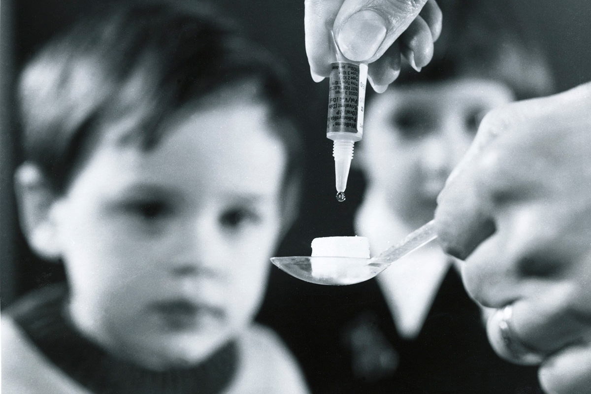 Löffel mit Würfelzucker auf den der Impfstoff getröpfelt wird. Im Hintergrund zwei Kinder.