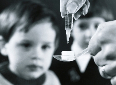 Löffel mit Würfelzucker auf den der Impfstoff getröpfelt wird. Im Hintergrund zwei Kinder.