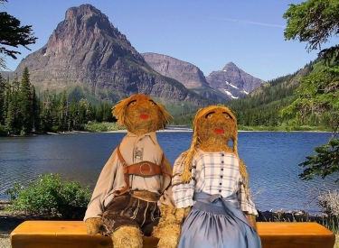Zwei Strohfiguren in Tracht auf einer Bank vor See und Bergen